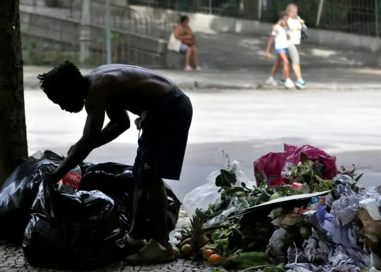 Foto de um homem negro na via pública. Ele está na calçada com corpo reclinado e mexendo com as mãos num saco de lixo. Veste bermuda preta, sapato marrom e está sem camisa.
