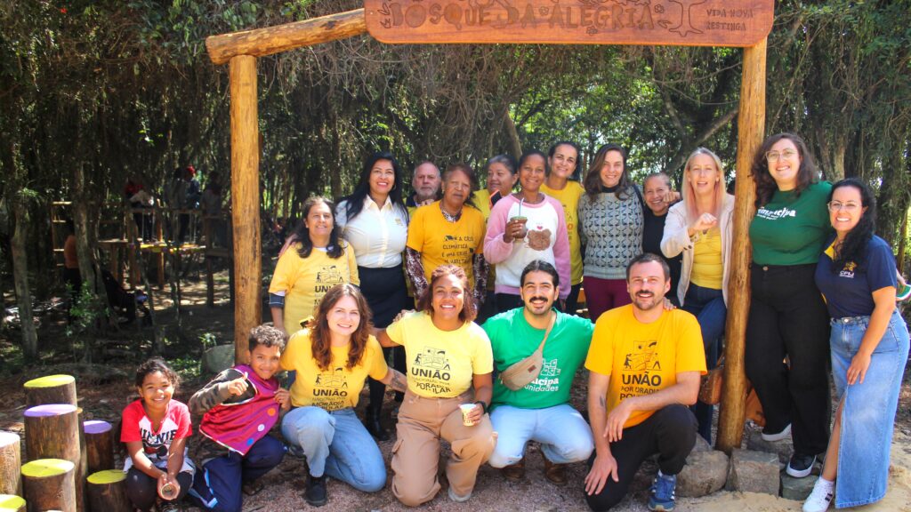Foto de grupo de mulheres, homens e crianças reunidos na praça Bosque da Alegria, em Porto Alegre. Estão sob uma placa de madeira que identifica o local. 