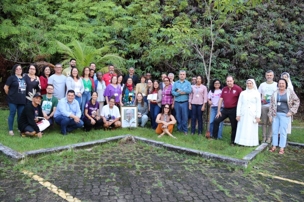 Foto de participantes do Encontro Nacional da Moradia e Favela, numa imagem com árvores ao fundo e no chão, na frente das pessoas, um retrato do Papa Francisco.