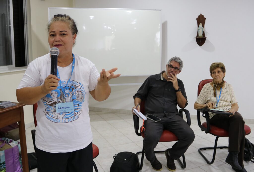 Foto de três pessoas participantes do Encontro de Moradia e Favela. À frente, de pé, com microfone na mão, está Leandra da Silva Ribeiro, moradora da ocupação Alcir Matos, em Manaus. Sentadas atrás dela estão outras duas pessoas participantes do evento.