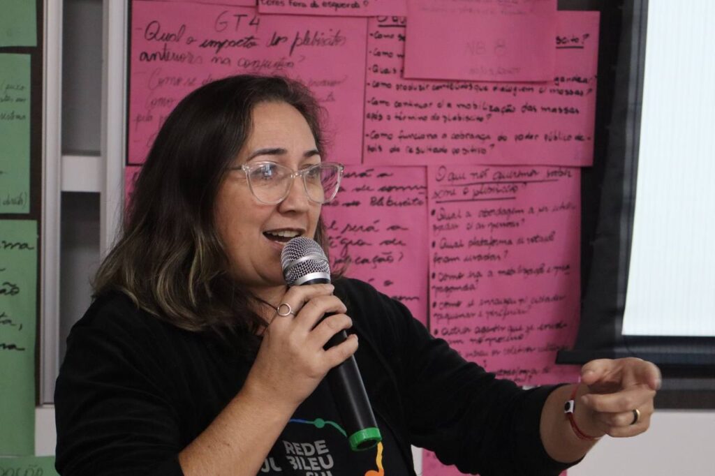 Foto de Rosilene Wansetto, secretária executiva do Jubileu Sul. Ela é branca, tem cabelos lisos castanhos, usa óculos e veste camiseta preta.