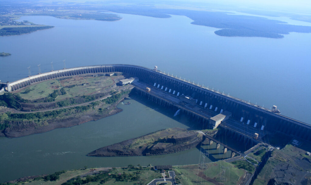 Foto da hidrelétrica, de Itaipu Binacional, com a usina na frente da imagem e atrás o grande reservatório de água represado.