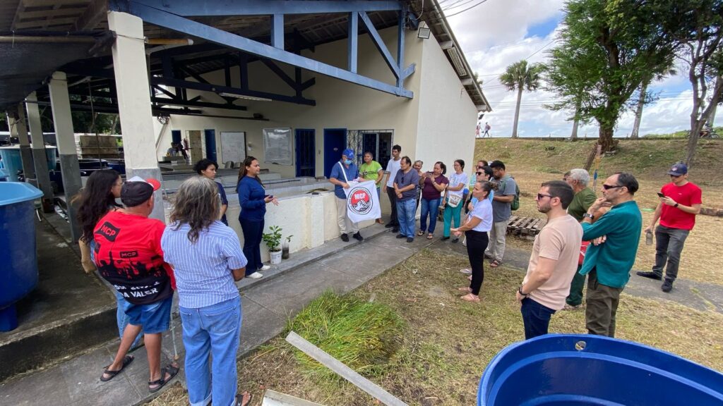 Grupo de cerca de 20 pessoas está reunido em frente a um prédio branco com estrutura azul na Universidade Federal do Ceará (UFC), durante visita técnica a laboratórios ligados ao projeto Sisteminha. À esquerda, tanques azuis de criação de peixes são visíveis. Uma mulher, ao centro, fala ao grupo; ao lado dela, um homem segura uma bandeira do Movimento dos Conselhos Populares (MCP). As pessoas presentes são lideranças comunitárias, moradores e integrantes de organizações sociais. Ao fundo, há árvores e céu parcialmente nublado. Foto: Ada Ponte.