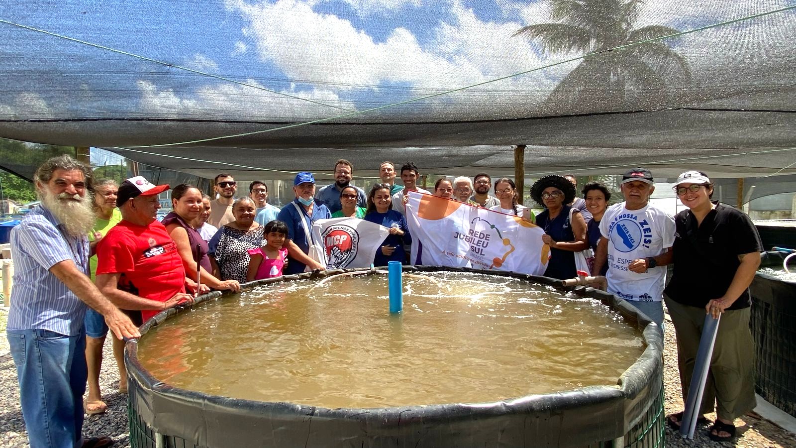 Grupo formado por cerca de 25 pessoas, incluindo moradores, lideranças comunitárias e integrantes de organizações sociais, posa em volta de um grande tanque circular de criação de peixes, durante visita técnica à Estação de Aquicultura da Universidade Federal do Ceará (UFC). As pessoas sorriem e seguram bandeiras do Movimento dos Conselhos Populares (MCP) e da Rede Jubileu Sul Brasil. A atividade ocorreu em ambiente coberto por tela de sombreamento, com céu azul visível ao fundo.