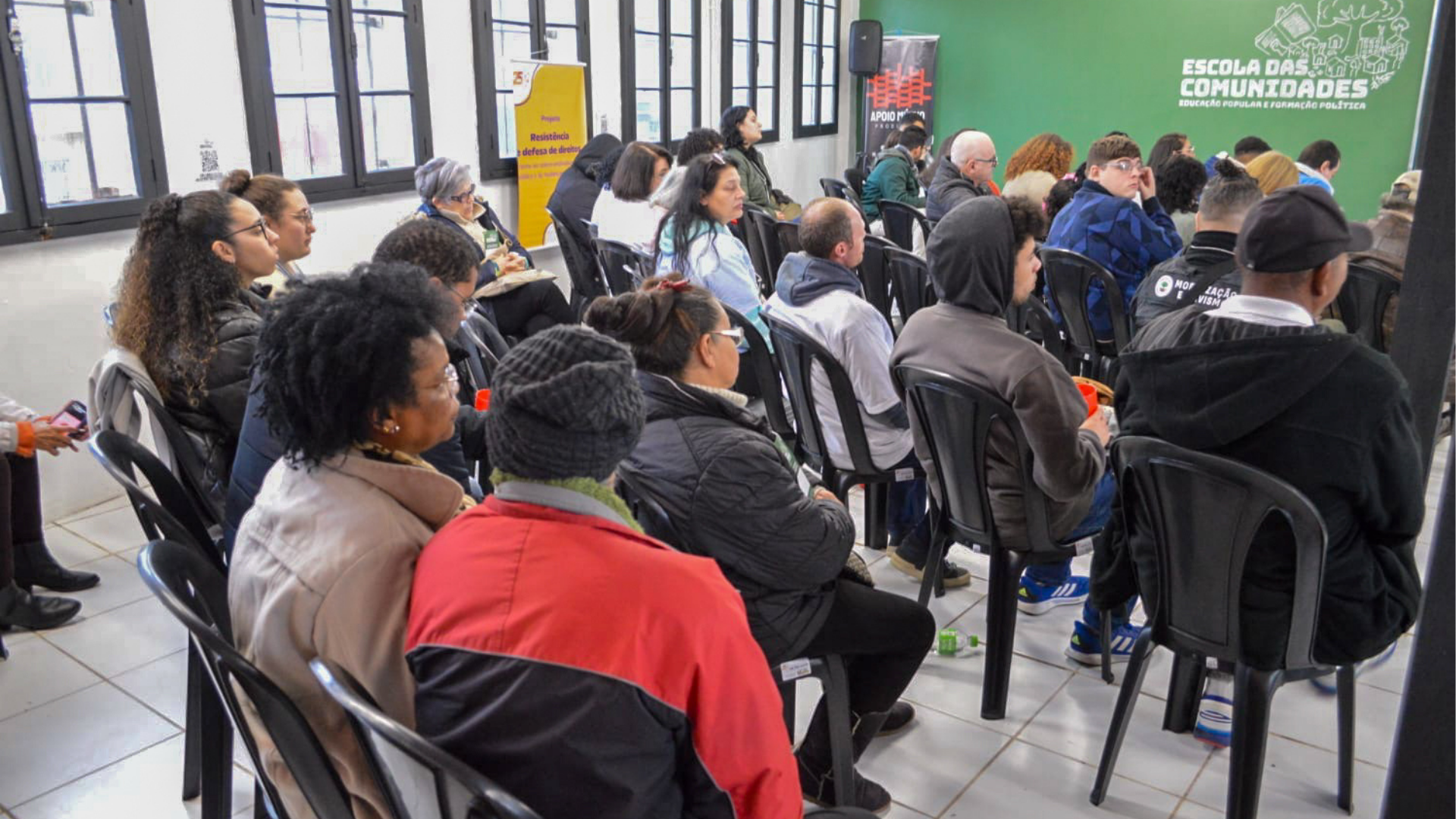 Sala cheia com cerca de 30 pessoas sentadas em cadeiras pretas de plástico, assistindo a uma atividade formativa. A maioria veste casacos e toucas, indicando clima frio. As pessoas estão voltadas para frente, com atenção à atividade. Na parede verde do fundo, o letreiro “Escola das Comunidades – Educação Popular e Formação Política” está visível, ao lado de banners com logomarcas de organizações populares.