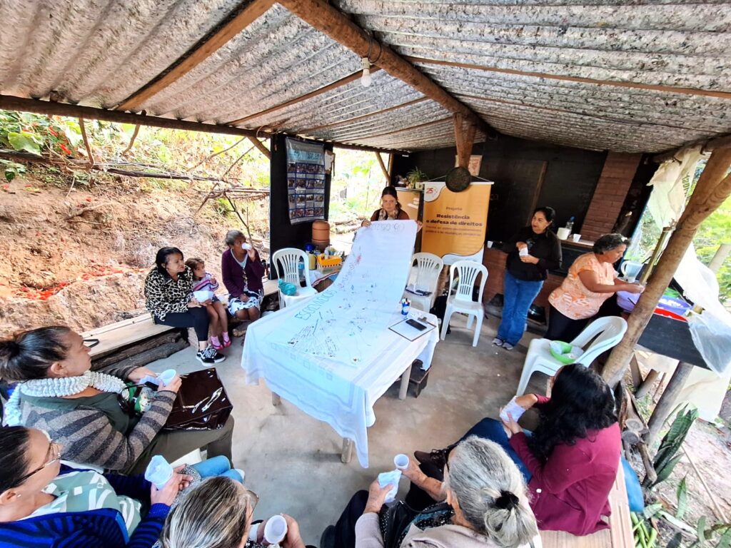 Mulheres de diferentes idades participam de uma roda de conversa em espaço coberto, ao ar livre, com vegetação ao fundo. Elas estão sentadas em círculo em torno de uma mesa com um grande tecido branco com registros escritos e desenhos, intitulado “Escrevivências Femininas”. Algumas participantes seguram copos ou cadernos, e há cartazes ao fundo, incluindo um com os dizeres “Projeto Resistência e Defesa de Direitos”. O ambiente é simples e acolhedor, com clima de escuta e troca de vivências. A atividade aconteceu em Belo Horizonte.