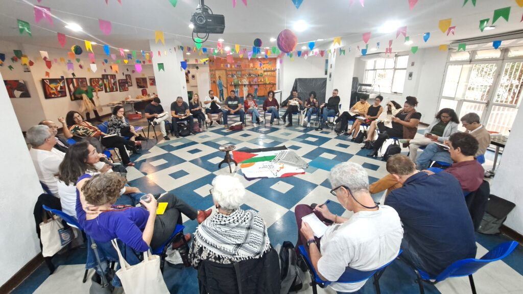 Foto de um encontro em formato de roda, com cerca de 40 pessoas sentadas em cadeiras azuis dispostas em círculo, em um salão decorado com bandeirinhas coloridas de festa junina. No centro da roda estão estendidas no chão duas bandeiras: uma da Palestina e outra da Rede Jubileu Sul Brasil com o mapa do Brasil e a frase ‘A vida acima da dívida’. A maioria dos participantes está atenta, tomando notas ou ouvindo atentamente. O ambiente tem chão quadriculado azul e branco, quadros coloridos nas paredes e boa iluminação natural
