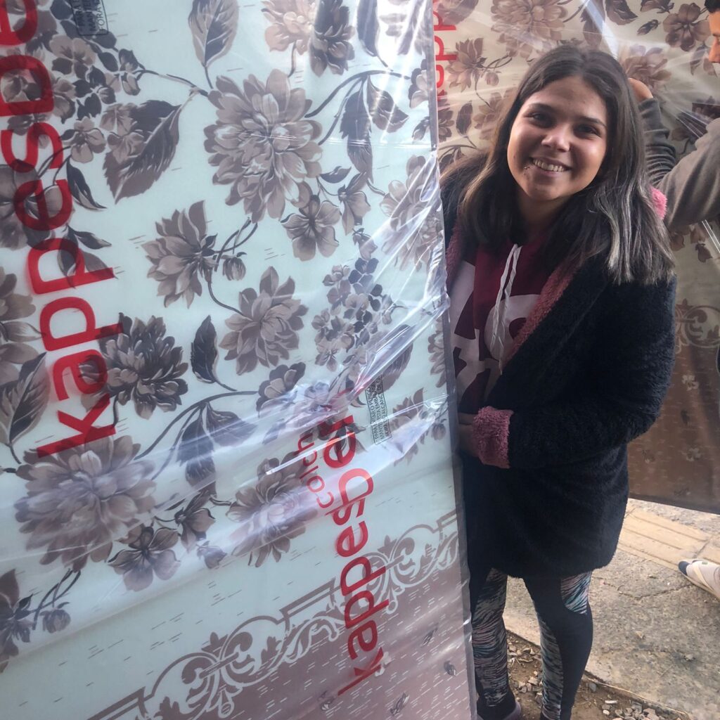 ma jovem sorridente segura um colchão novo ainda embalado em plástico transparente, estampado com flores em tons de marrom. Ela usa agasalho escuro e blusa clara, e está ao ar livre, possivelmente em área de entrega ou distribuição de donativos. A expressão da jovem transmite alegria e satisfação.