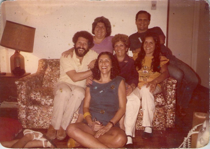 otografia colorida, de aparência antiga, retrata um grupo de sete pessoas sorridentes reunidas em uma sala de estar. Quatro pessoas estão sentadas em um sofá estampado e duas estão em pé atrás, com uma luminária de mesa acesa ao lado esquerdo. Uma mulher está sentada no chão à frente, com pernas cruzadas e expressão alegre. A imagem transmite um momento de união familiar e afeto. Ao fundo, parede clara e piso de madeira. A foto remete ao final dos anos 1970 ou início dos 1980.