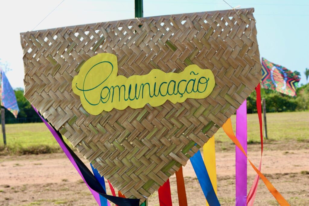 Placa de palha trançada em formato de coração invertido com a palavra “Comunicação” escrita em letra cursiva verde sobre um fundo amarelo. A placa está decorada com fitas coloridas penduradas nas laterais e aparece ao ar livre, com céu azul, vegetação e bandeiras coloridas ao fundo, indicando um ambiente festivo e comunitário.