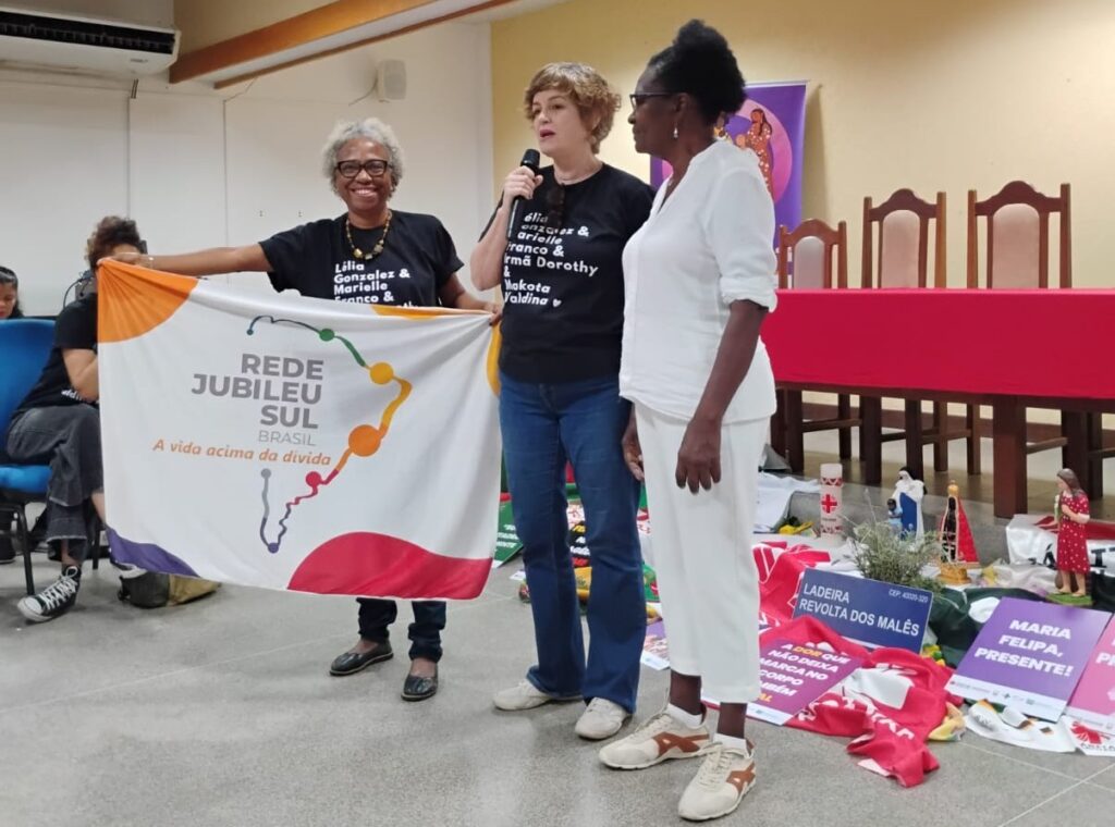 Três mulheres estão em pé no centro de uma sala durante um encontro. Duas delas são negras e vestem camisetas pretas com nomes de mulheres negras importantes; a terceira mulher é branca, segura um microfone e veste camiseta preta. As duas negras seguram uma bandeira da Rede Jubileu Sul Brasil com o lema "A vida acima da dívida". Ao fundo, uma mesa com pano vermelho e no chão há tecidos, cartazes e objetos simbólicos que remetem a lutas sociais e memoriais de mulheres. O ambiente transmite compromisso político, memória, mobilização e empoderamento feminino.