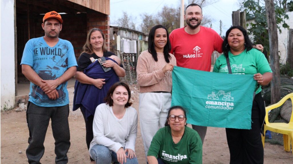 Sete pessoas, entre mulheres, um homem e um bebê, posam sorrindo para uma foto em uma comunidade do Rio Grande do Sul. Duas mulheres seguram uma bandeira verde com o logotipo “Emancipa Comunidades”. A cena acontece ao ar livre, em uma rua de terra, com casas simples ao fundo. O grupo demonstra união e engajamento em ações populares de base.