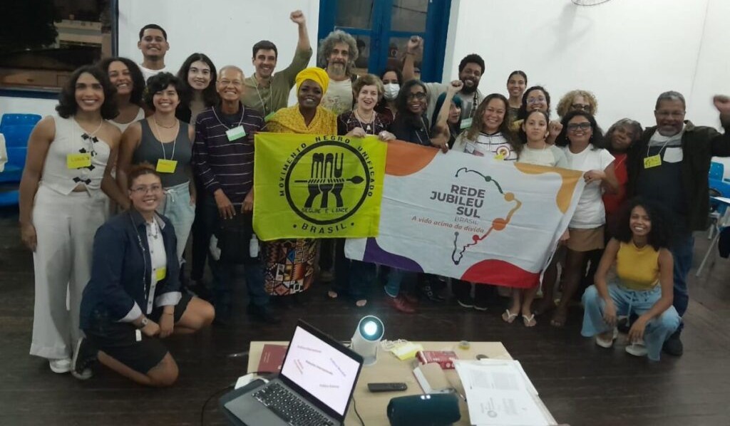 Grupo diverso de aproximadamente 25 pessoas reunidas em uma sala de aula, posando para foto coletiva. No centro, duas pessoas seguram bandeiras: uma do Movimento Negro Unificado (MNU) e outra da Rede Jubileu Sul Brasil, com o slogan “A vida acima da dívida”. Todas as pessoas estão sorrindo, algumas com o punho cerrado, em gesto de resistência. À frente, sobre uma mesa, há um notebook, projetor e materiais de apoio, indicando ambiente de formação ou encerramento de atividade educativa.