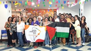 Grupo diverso de cerca de 30 pessoas reunidas sorrindo em uma sala decorada com bandeirolas coloridas, segurando duas bandeiras: uma da Rede Jubileu Sul Brasil, com o mapa do país estilizado e os dizeres 'A vida acima da dívida', e outra da Palestina, com faixas horizontais nas cores preta, branca e verde com um triângulo vermelho à esquerda. O ambiente é festivo, com chão quadriculado e clima de confraternização