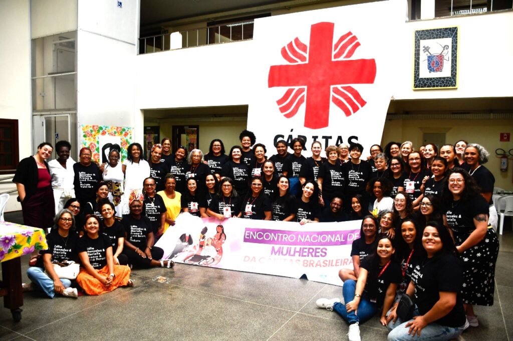 Grupo diverso de cerca de 50 mulheres posa sorrindo para uma foto coletiva no Encontro Nacional de Mulheres da Cáritas Brasileira. Elas estão reunidas em um espaço amplo e bem iluminado. A maioria veste camisetas pretas com dizeres em branco e segura uma faixa com os dizeres “Encontro Nacional de Mulheres da Cáritas Brasileira”. Ao fundo, um grande cartaz com o símbolo da Cáritas — uma cruz vermelha estilizada — está pendurado na parede. A imagem transmite união, força coletiva e protagonismo feminino.