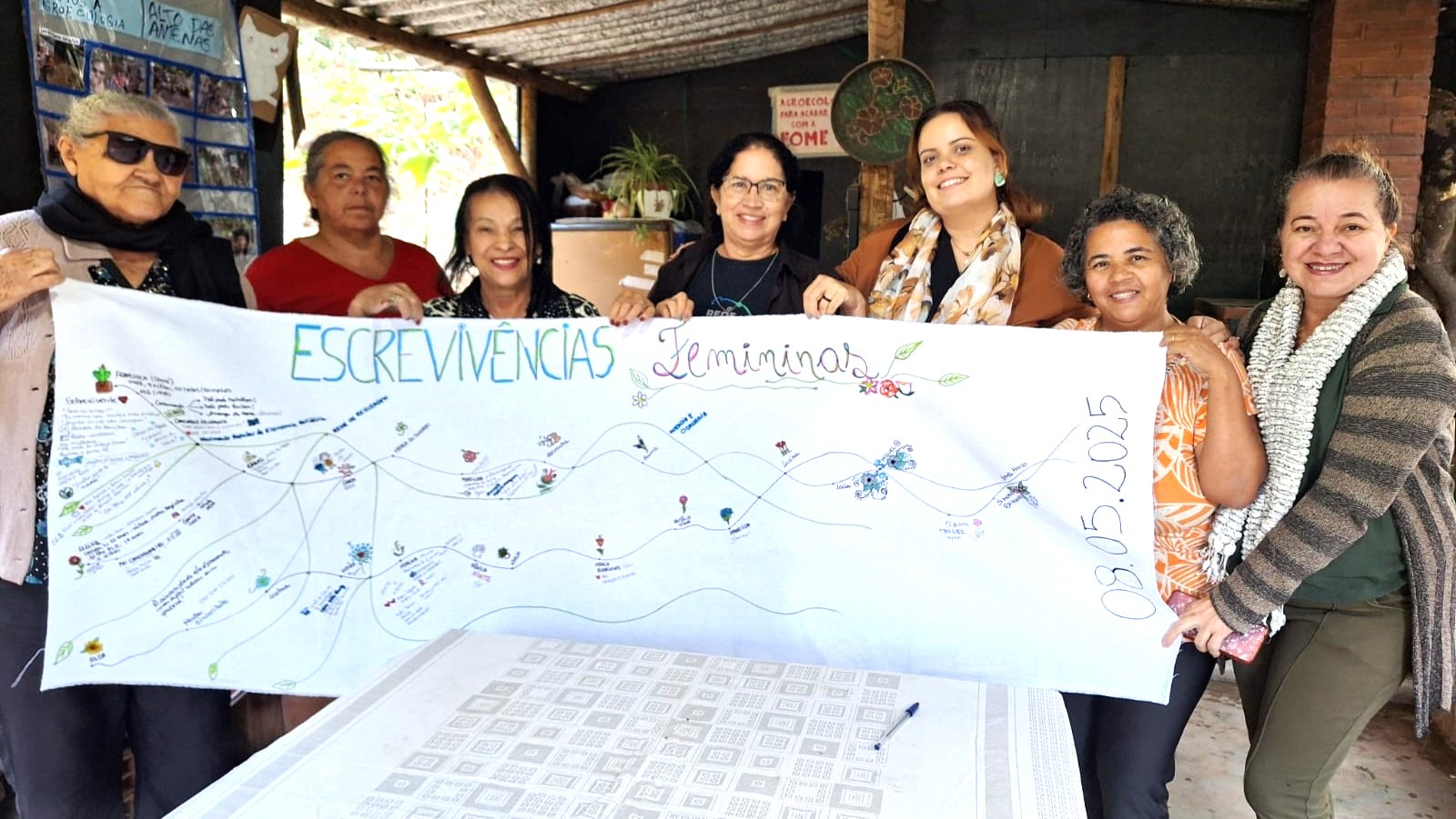 Oito mulheres sorridentes posam segurando um grande painel de tecido branco com a inscrição “Escrevivências Femininas” escrita à mão, acompanhada por desenhos de flores e linhas coloridas. O pano mostra registros textuais e gráficos feitos durante uma roda de conversa. A atividade aconteceu em Belo Horizonte, no dia 8 de maio de 2025, em ambiente rústico, com madeira, plantas e cartazes ao fundo. O encontro integra uma iniciativa de memória, escuta e partilha entre mulheres