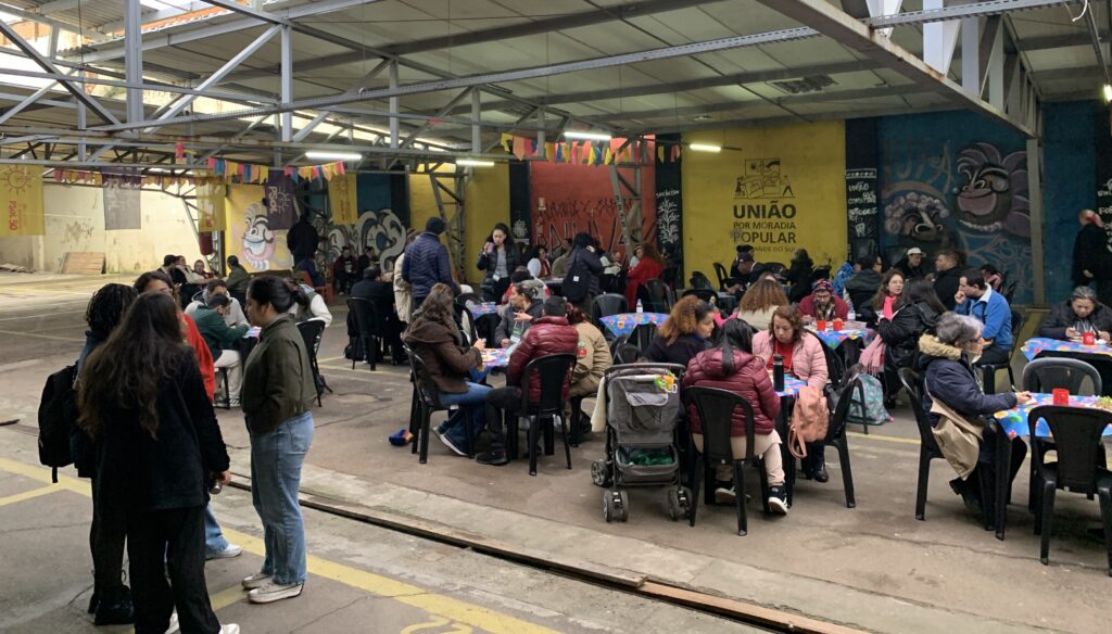 em um ambiente coberto e amplo, várias pessoas estão sentadas em cadeiras ao redor de mesas redondas com toalhas coloridas, participando de uma atividade comunitária com clima acolhedor. No fundo, há grafites nas paredes e um mural em amarelo com os dizeres “União por moradia popular”. Algumas pessoas estão em pé, conversando, e há uma atmosfera de encontro e diálogo. O espaço lembra um galpão adaptado para reunião de comunidade.