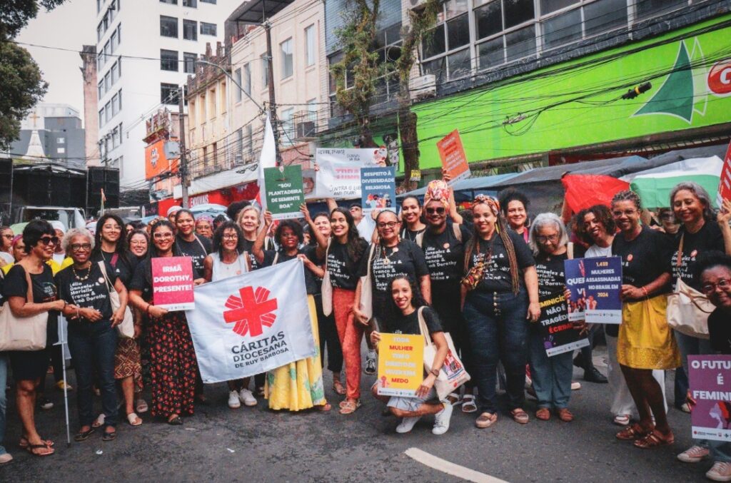 Grupo de cerca de 25 mulheres negras e algumas brancas reunidas em uma rua durante a Marcha das Mulheres Negras, parte da programação do Julho das Pretas em Salvador. Elas seguram cartazes com mensagens como “Irma Dorothy presente!” e “Respeito à diversidade religiosa”, além de um grande pano branco com o símbolo da Cáritas. Algumas vestem camisetas com nomes de mulheres negras importantes. A rua do centro da cidade está repleta de prédios comerciais ao fundo. A imagem transmite força, diversidade, ancestralidade e luta coletiva.
