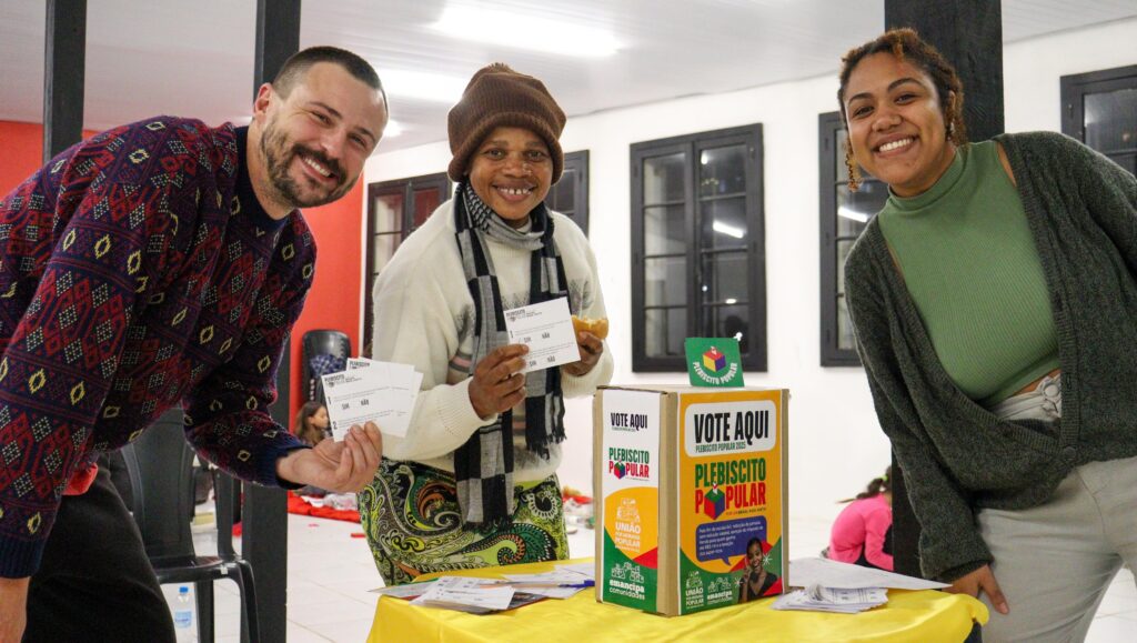 Três pessoas adultas — dois homens e uma mulher — sorriem para a câmera enquanto seguram cédulas de votação do Plebiscito Popular. Eles estão ao lado de uma urna simbólica colorida com os dizeres “Vote aqui – Plebiscito Popular 2025”. A mesa está coberta com um tecido amarelo e sobre ela há papéis organizados. O ambiente é uma sala clara, com janelas pretas e parede branca ao fundo. A atmosfera é de acolhimento e participação popular.