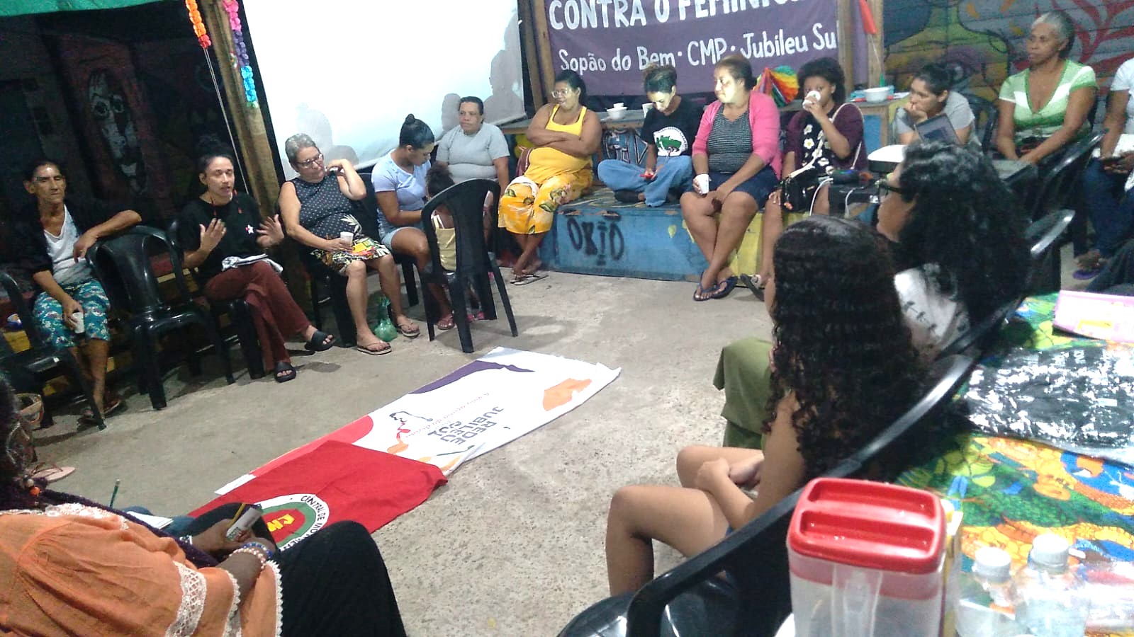 Mulheres e homens sentados em círculo participam de uma roda de conversa em um espaço comunitário. No centro, sobre o chão, estão estendidas duas bandeiras: uma da Rede Jubileu Sul Brasil e outra de movimento popular. Ao fundo, uma faixa roxa traz a frase “Contra o Fim do Mundo – Sopão do Bem CMP · Jubileu Sul”. O ambiente é simples e colorido, com murais pintados nas paredes, e as pessoas ouvem atentamente a fala de uma das participantes.