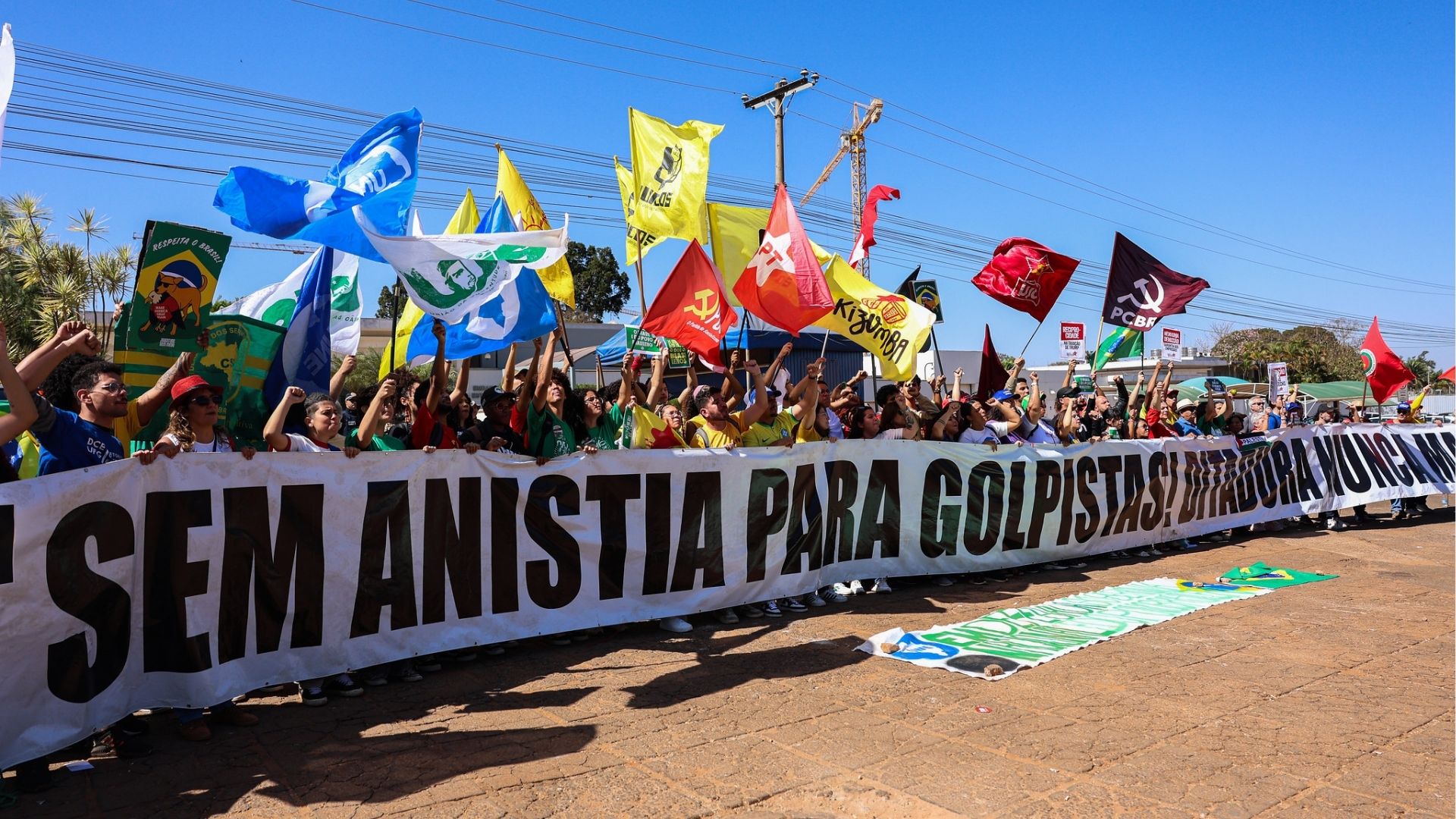 Manifestação com diversos manifestantes segurando bandeiras e uma faixa que diz 'Sem anistia para golpistas'. O protesto ocorre ao ar livre em um dia ensolarado, com participações de diferentes grupos políticos.