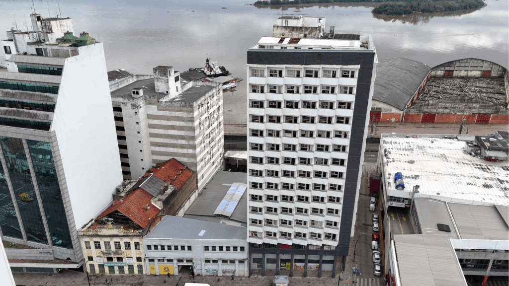 Imagem aérea mostra o centro de Porto Alegre (RS), com destaque para o Edifício Protetora, um prédio alto de fachada branca e janelas alinhadas, localizado próximo ao cais do rio Guaíba. Ao redor, aparecem outros edifícios comerciais e residenciais, além de galpões antigos próximos à margem do rio.