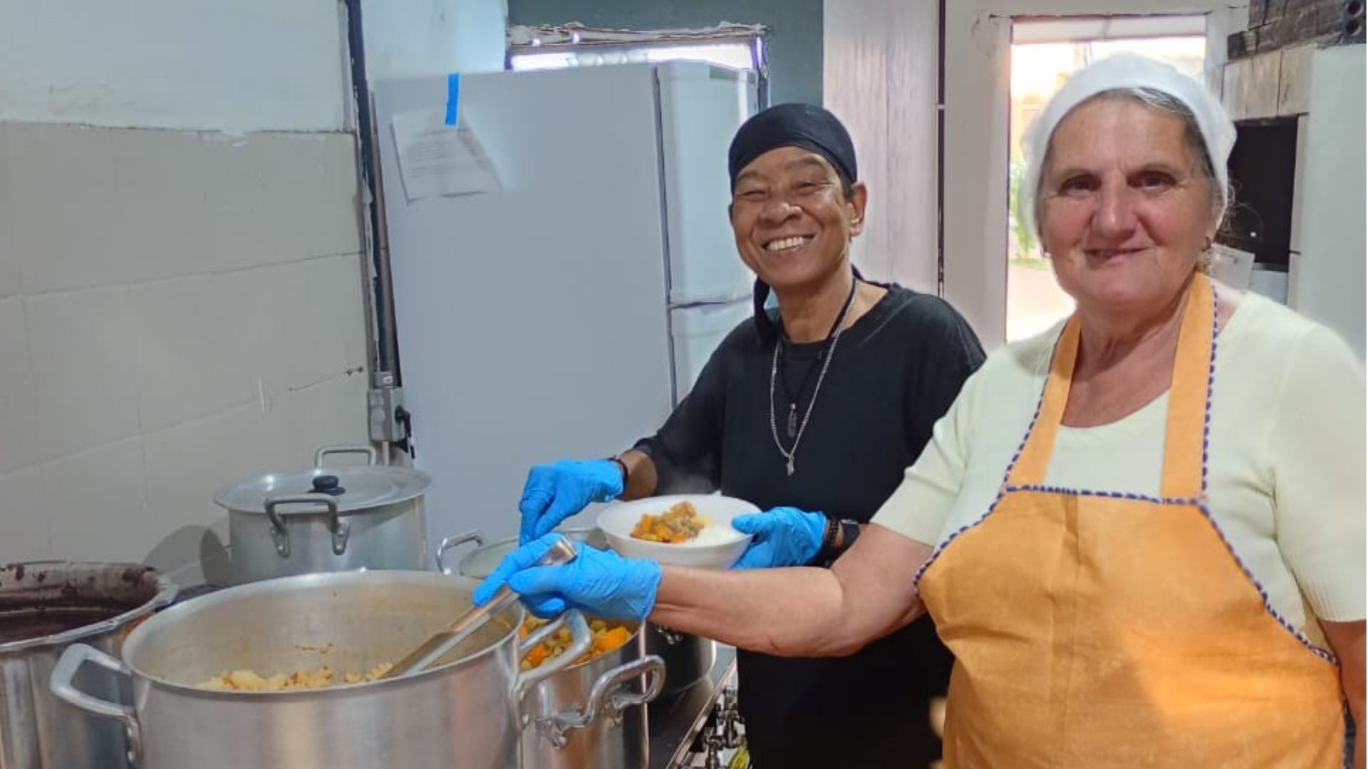 Duas pessoas estão em uma cozinha servindo comida em panelas grandes de inox. Um homem, sorridente, veste camiseta preta, bandana preta na cabeça e luvas azuis, segurando um prato de comida quente. Ao lado dele, uma mulher de avental laranja e blusa clara também usa touca e luvas, olhando para a câmera. Ambos estão em frente a panelas fumegantes sobre o fogão.