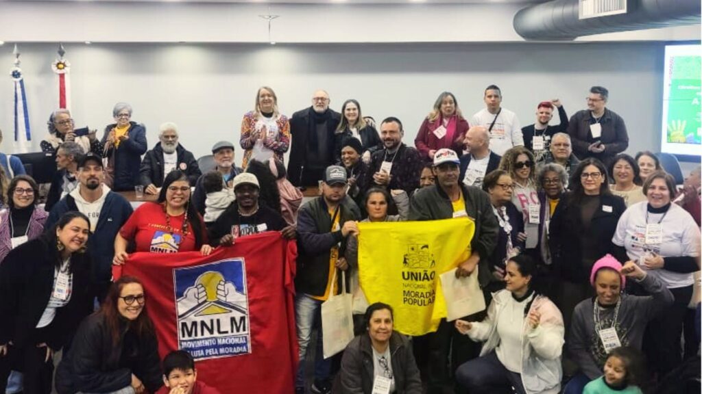 Grande grupo de pessoas reunidas em auditório, muitas delas em pé e outras sentadas ao centro. Alguns seguram faixas e banners de movimentos sociais (como MNLM e União Nacional por Moradia Popular). Ao fundo, detalhes da sala institucional com iluminação e telão. A cena exala mobilização, luta por direitos humanos e articulação coletiva