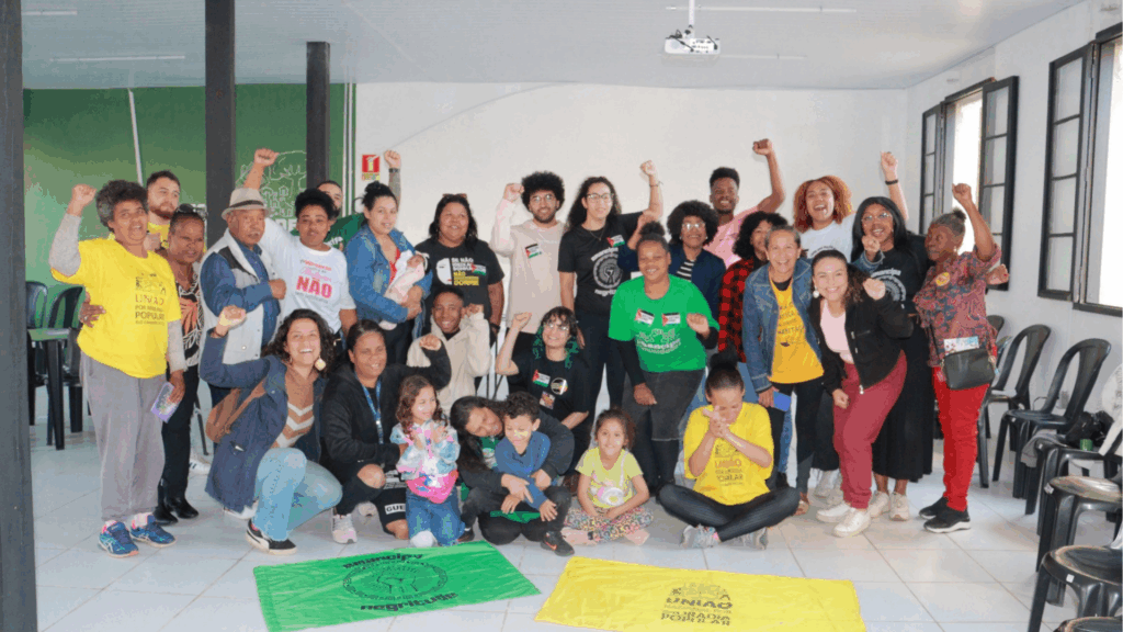 Moradoras e lideranças reunidas em sala, algumas com punhos erguidos em gesto de resistência, outras sorrindo; ao fundo, pintura verde na parede e cadeiras pretas. A cena transmite empoderamento coletivo, debate social e engajamento de mulheres em defesa de direitos.