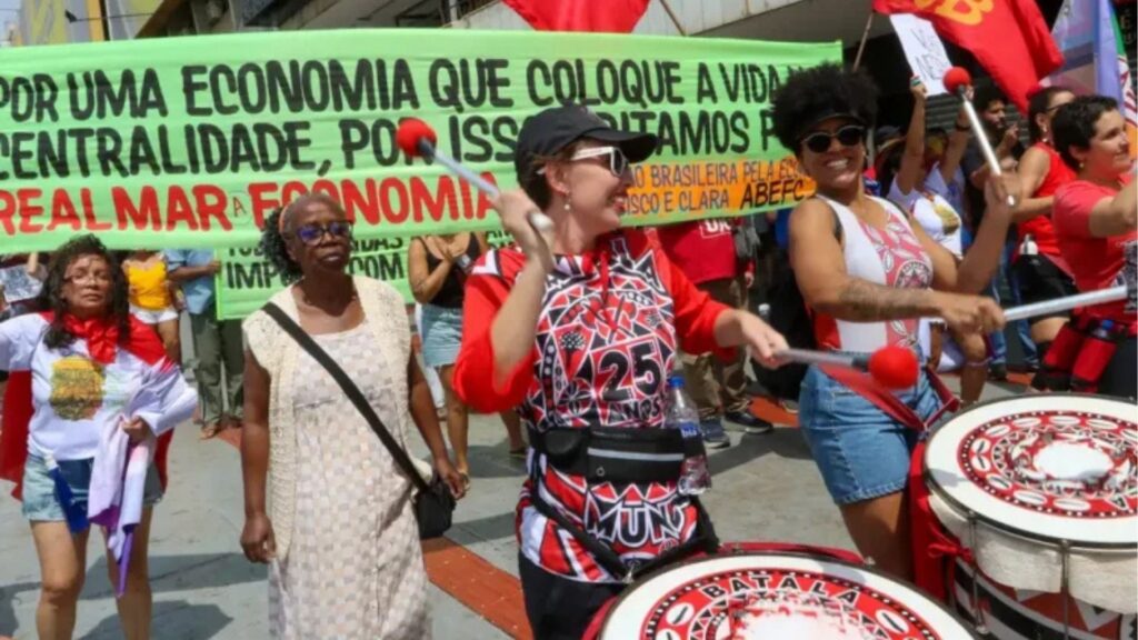 Manifestação popular em uma rua no Brasil. No centro, duas mulheres tocam grandes tambores decorados de vermelho, branco e preto, sorrindo e marchando. Ao fundo, diversas pessoas seguram uma faixa verde com letras pretas e vermelhas, onde se lê: “Por uma economia que coloque a vida na centralidade, por isso gritamos por...”. Ao redor, participantes de movimentos sociais carregam bandeiras vermelhas e caminham em conjunto. A cena expressa luta, música e reivindicação por justiça social e econômica.
