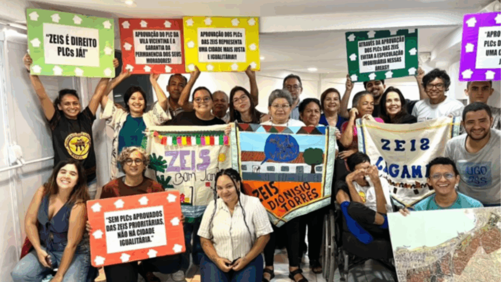 Um grupo diverso de pessoas posa sorridente em ambiente interno, segurando cartazes coloridos e faixas que celebram a aprovação dos Projetos de Lei Complementares (PLCs) das Zonas Especiais de Interesse Social (ZEIS) de Fortaleza. Os cartazes trazem frases como “ZEIS é direito, PLCs já!”, “Aprovação dos PLCs das ZEIS representa uma cidade mais justa e igualitária!” e “Sem PLCs aprovados das ZEIS prioritárias, não há cidade igualitária”. Na frente do grupo, também são exibidas bandeiras artesanais dos territórios ZEIS Bom Jardim, ZEIS Dionísio Torres e ZEIS Lagamar, representando a luta coletiva pelo direito à moradia e à cidade.