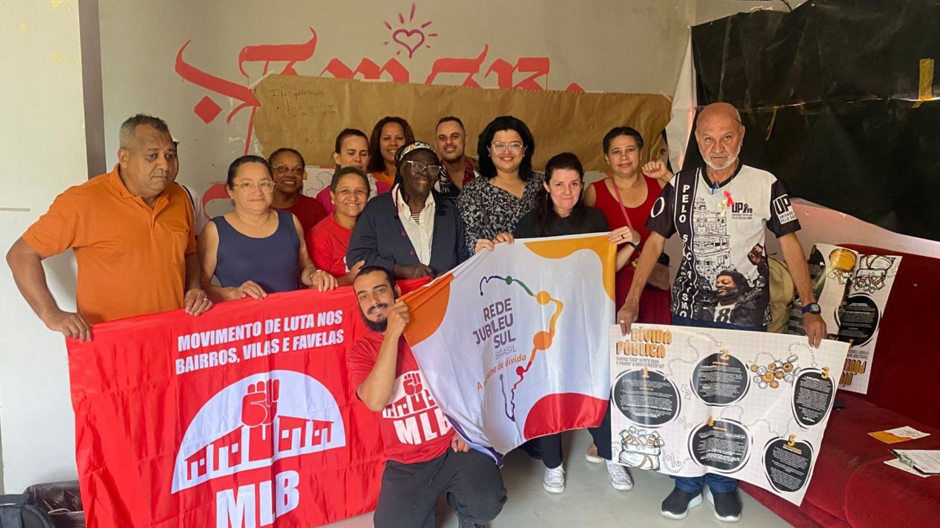 Grupo de pessoas reunidas em uma sala durante o encerramento do curso sobre dívida pública promovido pela Rede Jubileu Sul Brasil em parceria com o Movimento de Luta nos Bairros, Vilas e Favelas (MLB). Elas posam sorrindo e seguram bandeiras das duas organizações e cartazes informativos sobre dívida pública. Ao fundo, há um cartaz pardo e uma parede com inscrições em vermelho.
