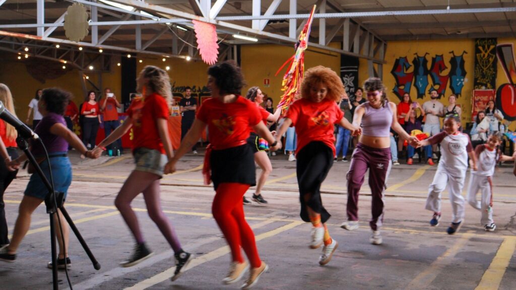 Grupo de pessoas dançando em roda, segurando mãos e sorrindo num espaço urbano coberto. Algumas usam camisetas vermelhas e roupas coloridas; ao fundo, grafites e observadores. O ambiente transmite festa, expressão cultural e união popular
