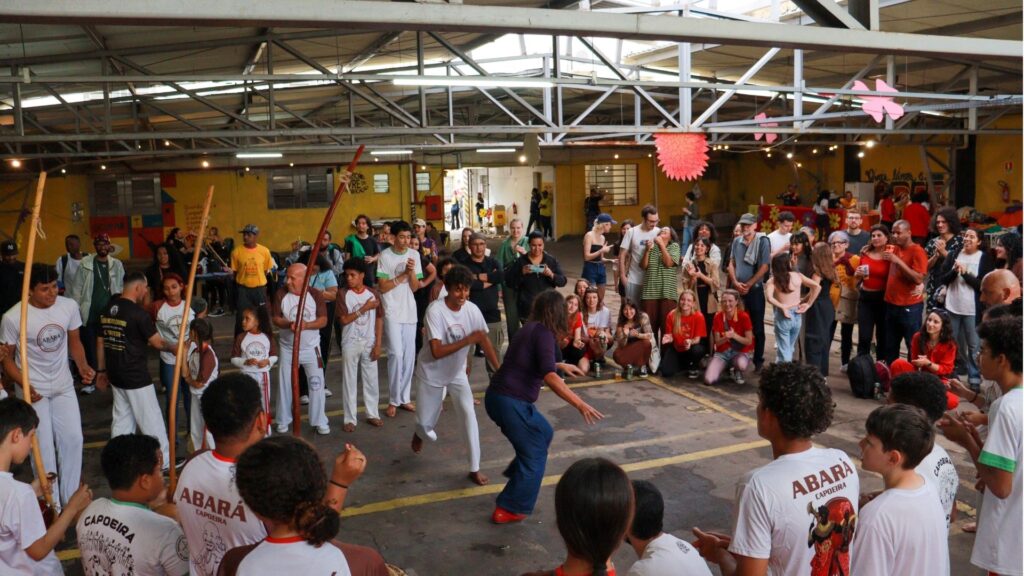 Roda de capoeira realizada em um espaço coberto, com praticantes (crianças, jovens e adultos) posicionados em círculo e observadores ao redor. No centro, dois participantes dançam e trocam golpes de capoeira. Ao fundo, público aplaude, fotografa e interage com entusiasmo. A cena transmite cultura popular, movimento, celebração comunitária e energia.