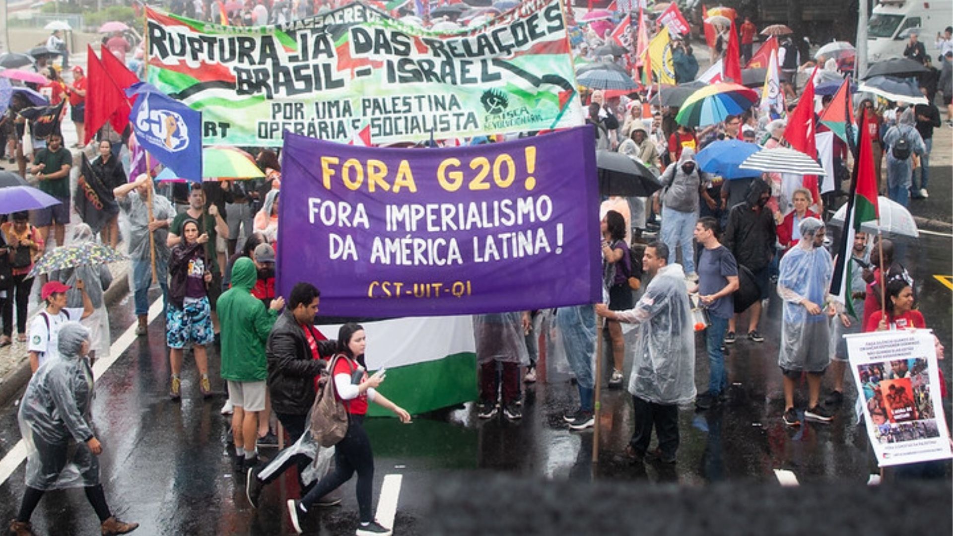Manifestação de rua sob chuva com dezenas de pessoas usando capas plásticas e segurando guarda-chuvas. No centro, manifestantes carregam uma grande faixa roxa com os dizeres “Fora G20! Fora imperialismo da América Latina!”. Ao fundo, outra faixa pede a ruptura das relações entre Brasil e Israel e defende uma Palestina operária e socialista. Diversas bandeiras e cartazes coloridos aparecem, compondo um ato de protesto coletivo. Foto de Rodrigo Matos.