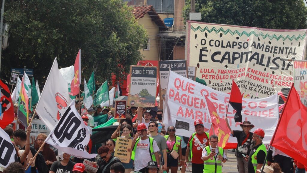 A imagem mostra uma grande marcha de rua com diversas pessoas segurando bandeiras, faixas e cartazes. Entre as mensagens visíveis estão “O colapso ambiental é capitalista”, “Agro é fogo! Acabar com o agronegócio para poder respirar” e “Que os ricos paguem pela crise ambiental”. Há bandeiras de movimentos sociais, sindicatos e organizações populares. As pessoas marcham sob sol forte, ocupando a via, com árvores e prédios ao fundo, em um ato de denúncia contra o capitalismo, o agronegócio e as falsas soluções climáticas.