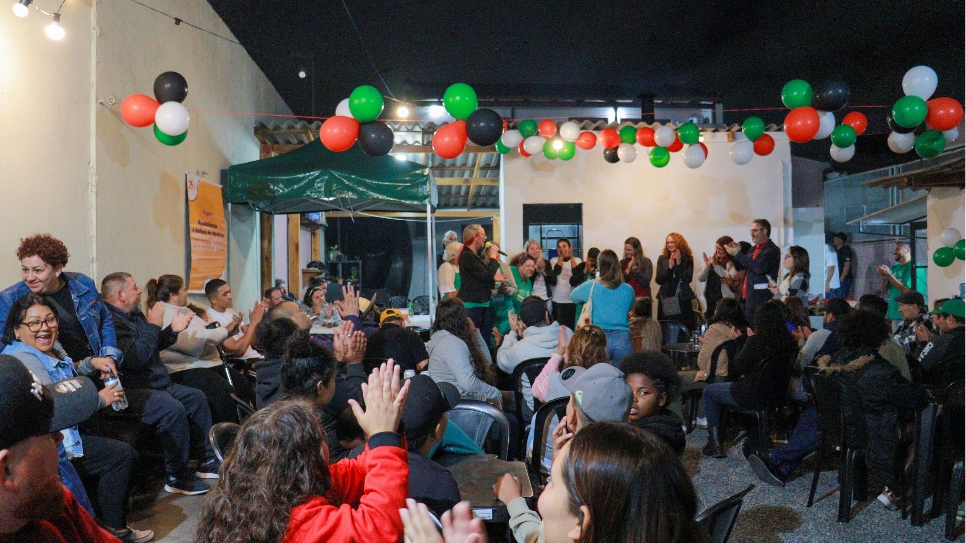 Grupo de pessoas participa de um evento comunitário à noite em um espaço decorado com balões nas cores verde, vermelho, branco e preto. As pessoas estão sentadas e aplaudem enquanto um grupo se apresenta em frente ao público, sob uma tenda verde. O ambiente é alegre e acolhedor, representando uma ação coletiva e solidária.