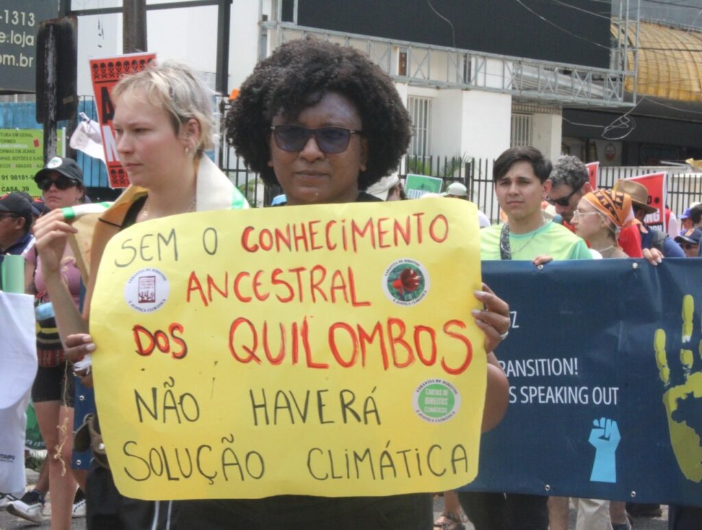 Uma pessoa em meio a uma marcha segura um cartaz amarelo escrito “Sem o conhecimento ancestral dos quilombos não haverá solução climática”. Ela usa óculos escuros e está cercada por outras pessoas manifestando, algumas carregando faixas e cartazes. Ao fundo, vê-se a rua, prédios comerciais e participantes da mobilização, sugerindo um ato público relacionado à justiça climática.