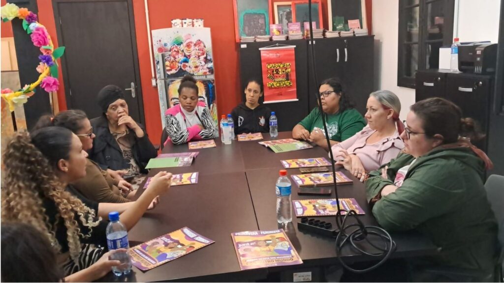 Grupo de mulheres participa de uma roda de conversa em uma sala colorida, com paredes vermelhas e decoração com flores de papel. Elas estão sentadas ao redor de uma mesa retangular, conversando e segurando cartilhas com o título “Palavra Feminista”. Sobre a mesa há garrafas de água e materiais impressos. O ambiente transmite um clima de diálogo e troca de experiências sobre temas de luta e organização das mulheres.