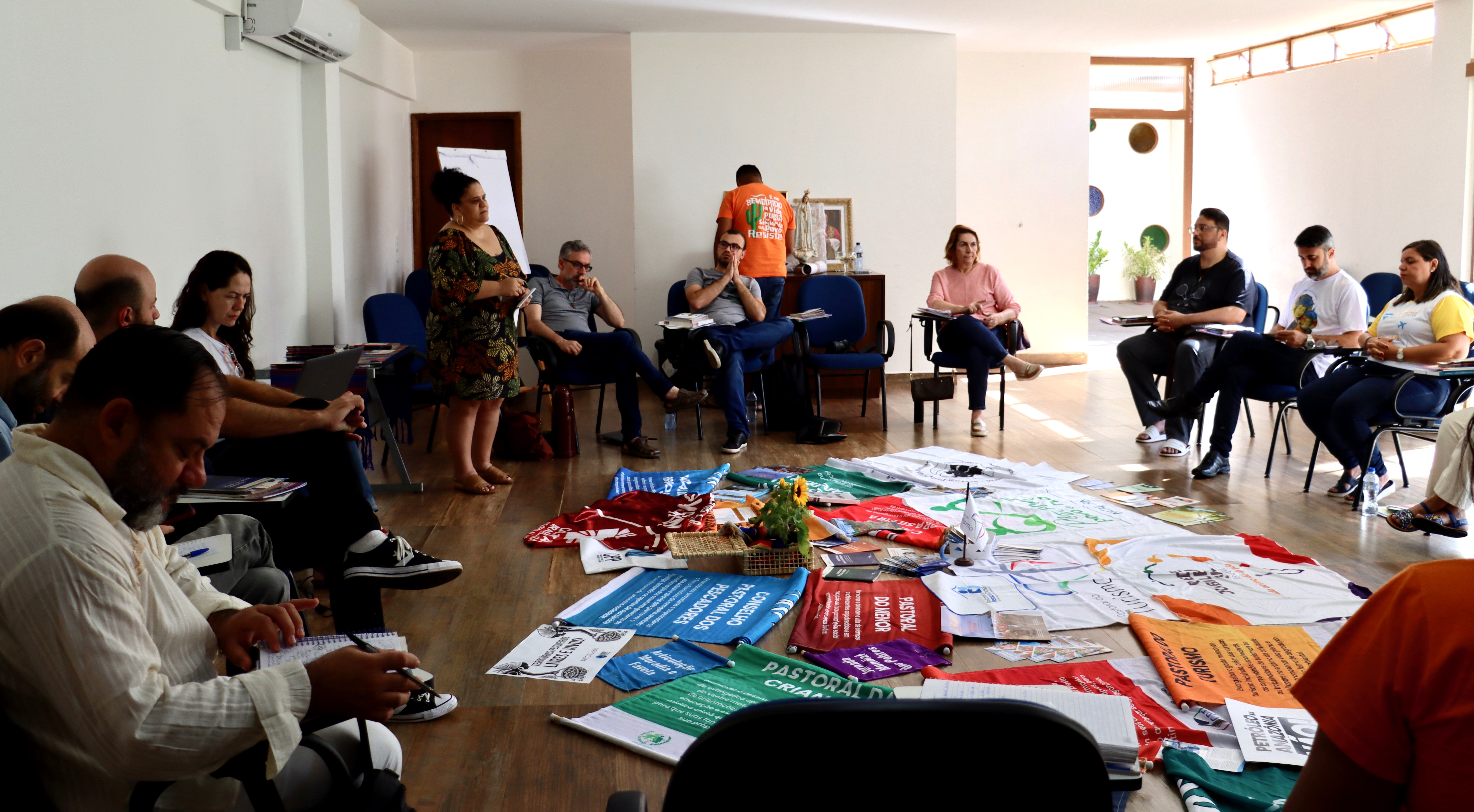 Participantes do Encontro de Comunicação Sócio-Transformadora estão reunidos em círculo em uma sala ampla e iluminada, sentados em cadeiras. No centro do espaço, há faixas, cartazes e materiais de diversas pastorais e organizações sociais dispostos no chão, como Pastoral da Criança, Pastoral Carcerária, Cáritas e Jubileu Sul. Uma mulher está em pé, conduzindo a fala, enquanto outras pessoas escutam, anotam e refletem coletivamente. A cena expressa diálogo, planejamento conjunto e construção coletiva de estratégias de comunicação popular.