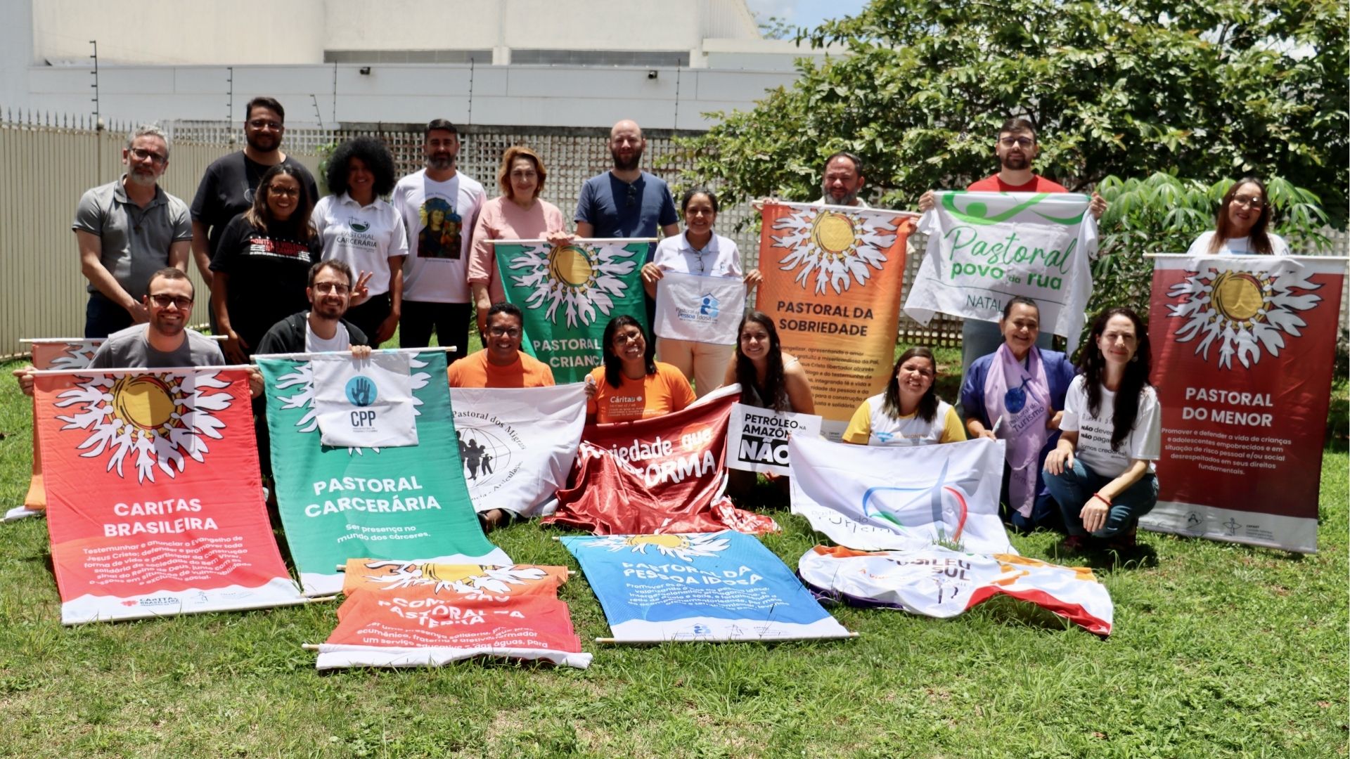 Grupo diverso de cerca de 25 pessoas posa ao ar livre, em um gramado, durante o Encontro de Comunicação Sócio-Transformadora da Cepast/CNBB. As participantes seguram faixas e bandeiras de diferentes pastorais e organizações sociais, como Cáritas Brasileira, Pastoral Carcerária, Pastoral do Menor, Pastoral da Criança, Pastoral da Pessoa Idosa, Pastoral da Sobriedade, Pastoral do Povo da Rua, CPP e Jubileu Sul. Ao fundo, há árvores e um prédio claro. A imagem transmite articulação em rede, diversidade e compromisso coletivo com justiça social e direitos humanos.