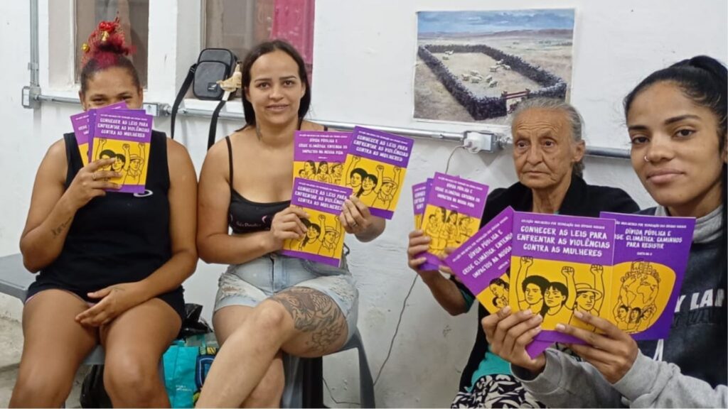 Quatro mulheres sentadas lado a lado, segurando e exibindo várias cartilhas de capa roxa e amarela com ilustrações e o título “Conhecer as leis para enfrentar as violências contra as mulheres”. Três delas são jovens e uma é uma senhora idosa. Todas olham para a câmera; uma das mulheres mais jovens sorri, outra cobre parcialmente o rosto com as cartilhas. A cena ocorre em um ambiente simples, com paredes claras e uma fotografia pendurada ao fundo. A atmosfera transmite orgulho, participação e fortalecimento comunitário.