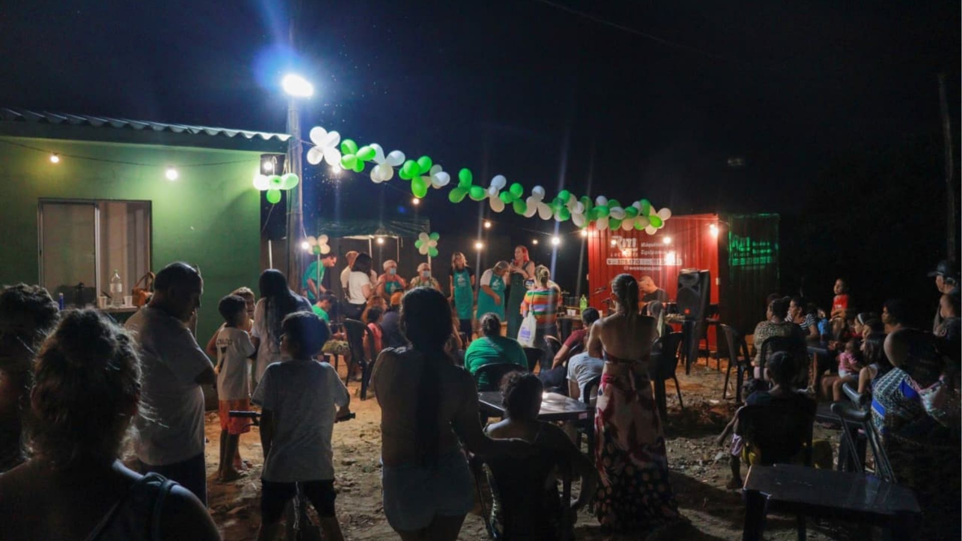 Cena noturna de uma atividade comunitária ao ar livre. Diversas pessoas, incluindo mulheres, homens e crianças, estão reunidas em frente a um palco simples decorado com balões verdes e brancos. Algumas mulheres no palco usam aventais verdes e seguram microfones, parecendo conduzir falas ou apresentações. O espaço é iluminado por lâmpadas penduradas e refletores, com mesas e cadeiras ocupadas pelo público. Ao fundo, há estruturas como um contêiner vermelho e uma casa verde. A atmosfera é de convivência, celebração e participação coletiva.