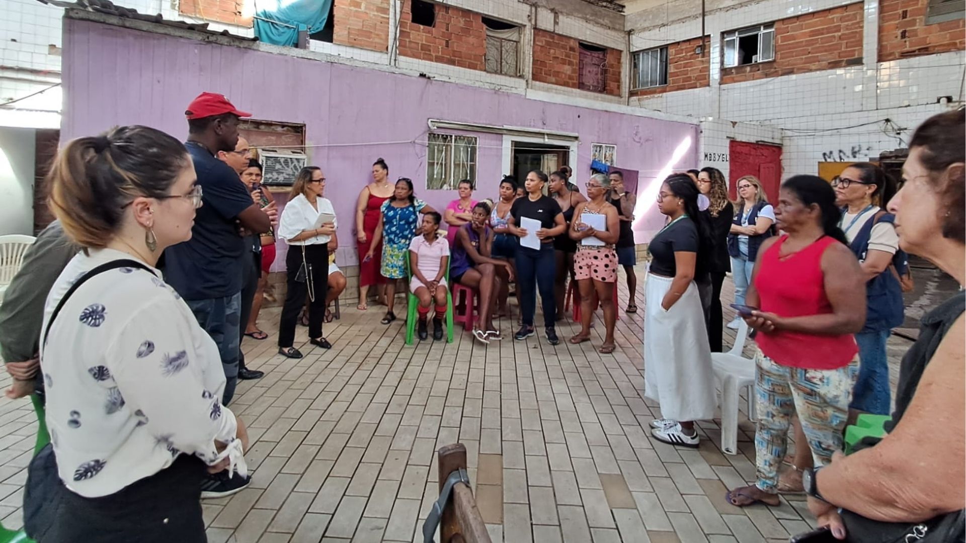 Um grupo diverso de mulheres e alguns homens participa de uma roda de conversa em um pátio interno de uma ocupação ou comunidade. As pessoas estão em pé ou sentadas em cadeiras e banquinhos, formando um círculo diante de uma parede roxa. Muitas seguram cadernos ou papéis, indicando uma atividade formativa ou reunião comunitária. O ambiente é simples, com paredes de alvenaria exposta e janelas sem vidros. A cena transmite escuta coletiva, organização comunitária e troca de experiências.