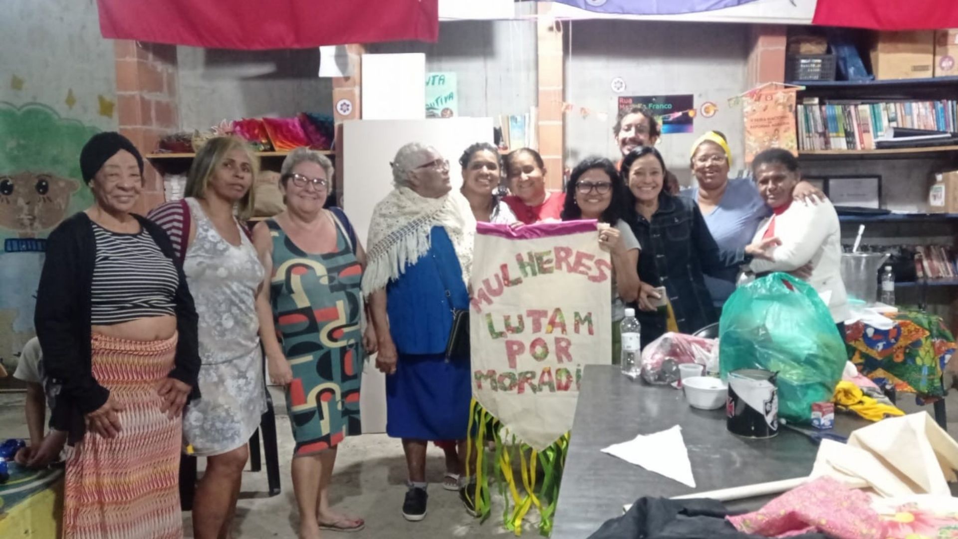 Grupo de mulheres de diferentes idades reunidas em espaço comunitário interno, sorrindo para a foto. Ao centro, seguram um cartaz artesanal com a frase “Mulheres lutam por moradia”. O ambiente tem estantes com livros e materiais ao fundo, além de mesa com objetos e garrafas.