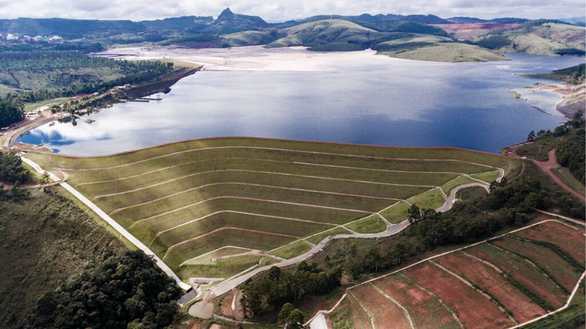 Vista aérea de uma barragem com taludes em degraus cobertos por grama, formando linhas horizontais ao longo da encosta. À frente, há uma estrada de acesso e áreas de vegetação. Ao fundo, um grande reservatório de água se estende até colinas verdes e montanhas, sob céu parcialmente nublado. À direita, aparecem áreas de solo avermelhado exposto.