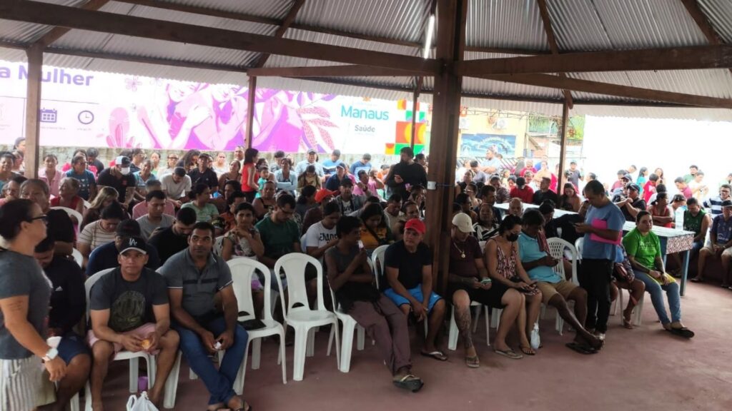 Dezenas de moradores estão reunidos sob uma estrutura coberta com vigas de madeira e telhado metálico, sentados em cadeiras plásticas brancas e aguardando atendimento. Algumas pessoas estão em pé ao fundo, formando fila. Ao fundo da imagem, há um painel com a inscrição “Manaus Saúde” e ilustrações em tons de rosa. O ambiente indica uma ação comunitária ou cadastramento coletivo, com grande participação da população local.
