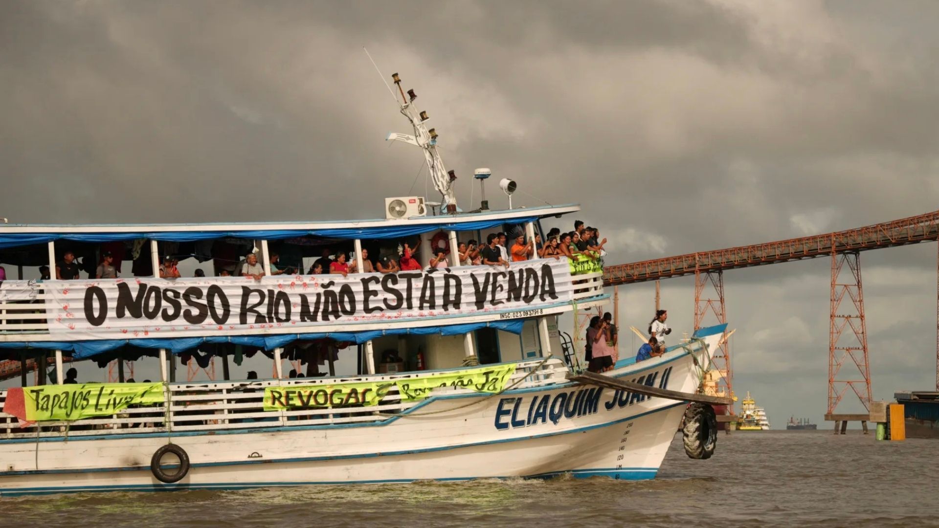 Embarcação no rio com dezenas de pessoas a bordo segurando faixas de protesto. A maior delas diz “O nosso rio não está à venda”. Outras faixas trazem mensagens como “Tapajós Livre” e “Revogar já”. Ao fundo, aparece uma grande estrutura metálica usada para transporte de grãos sobre o rio, sob céu nublado.