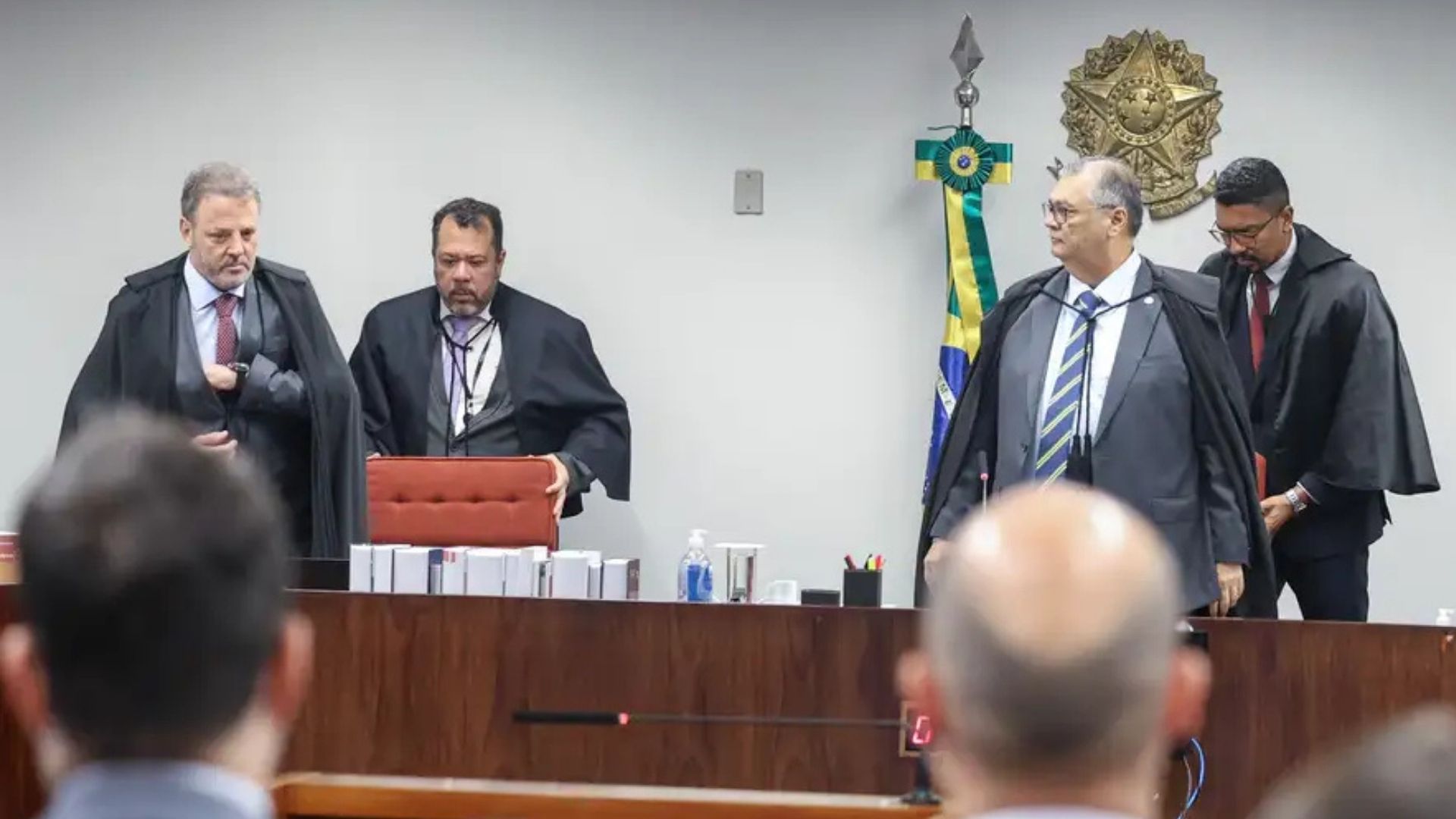 Quatro homens vestindo ternos escuros e togas estão em um plenário de tribunal. Três deles estão atrás de uma mesa com diversos volumes de processos organizados, enquanto outro permanece um pouco afastado. Ao fundo, aparecem a bandeira do Brasil e o brasão da República fixado na parede. Em primeiro plano, vê-se a parte de trás da cabeça de pessoas sentadas, acompanhando a sessão.
