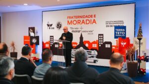 Um bispo católico, vestido de preto e usando óculos, fala ao microfone em um púlpito transparente durante um evento em auditório. Ao fundo, um grande painel com o tema “Fraternidade e Moradia” traz a frase “Ele veio morar entre nós” (João 1,14), além das logomarcas da CNBB e da Campanha da Fraternidade. Também aparece a data da Coleta Nacional da Solidariedade (29 de março) e a expressão “Terra, Teto, Trabalho”. Na plateia, várias pessoas sentadas acompanham a fala. A decoração inclui ilustrações de casas, prédios e uma igreja, remetendo ao tema da moradia, além de uma imagem de Nossa Senhora em destaque lateral.